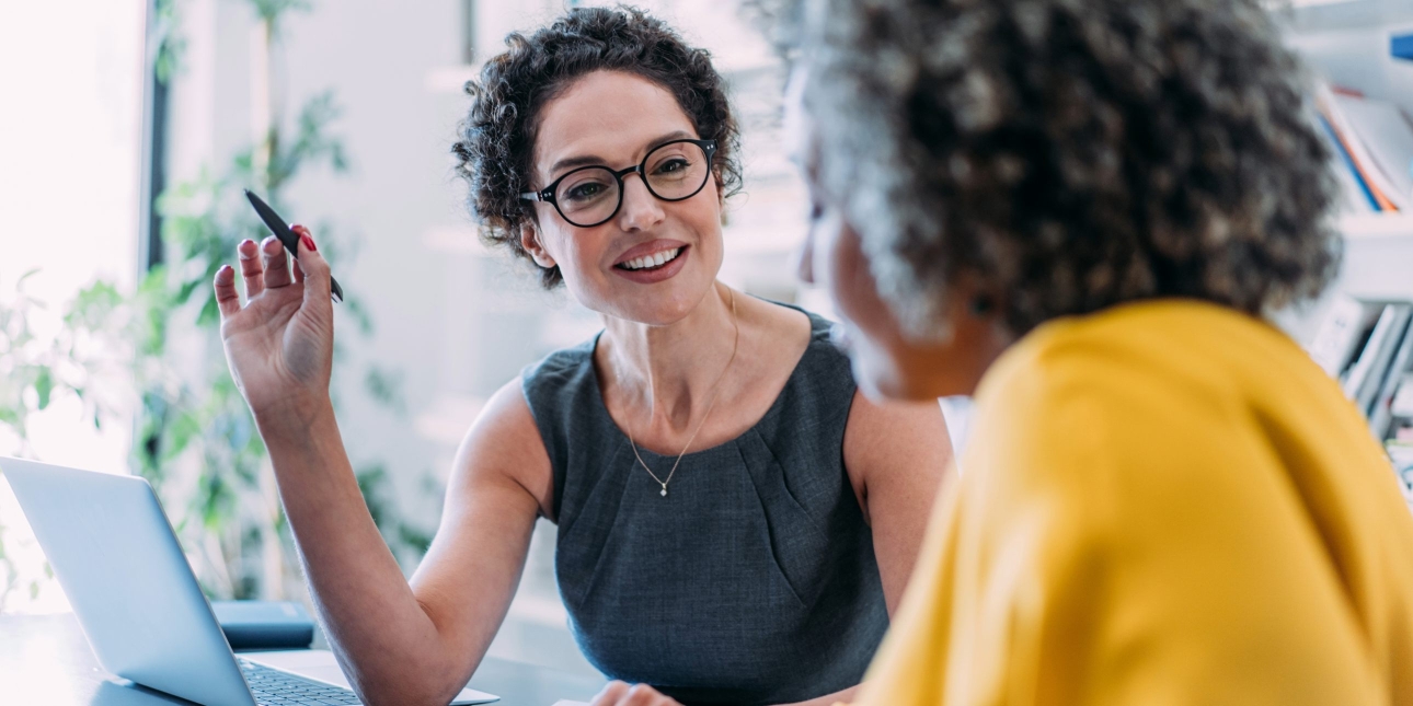 A white woman with short dark hair, weatring a grey top and glasses is holding a pen and sitting at a laptop is talking to a woman who is side on to the camera and wearing a yello top